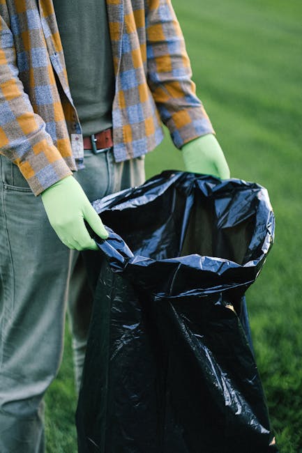 A person wearing a casual plaid shirt with shades of yellow, blue, and white, along with a green long-sleeved top underneath, holds open a large black plastic rubbish bag lined with silver interior li