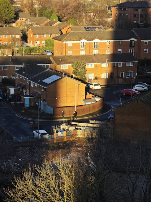 An aerial view of a small residential area featuring a roundabout surrounded by low-rise brick and wooden buildings, with parked cars along the streets and in parking spaces. The scene includes a mix 